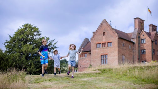 3 children holding hands run down the hill in the parkland at Chartwell. The red brick house and trees are in the background.